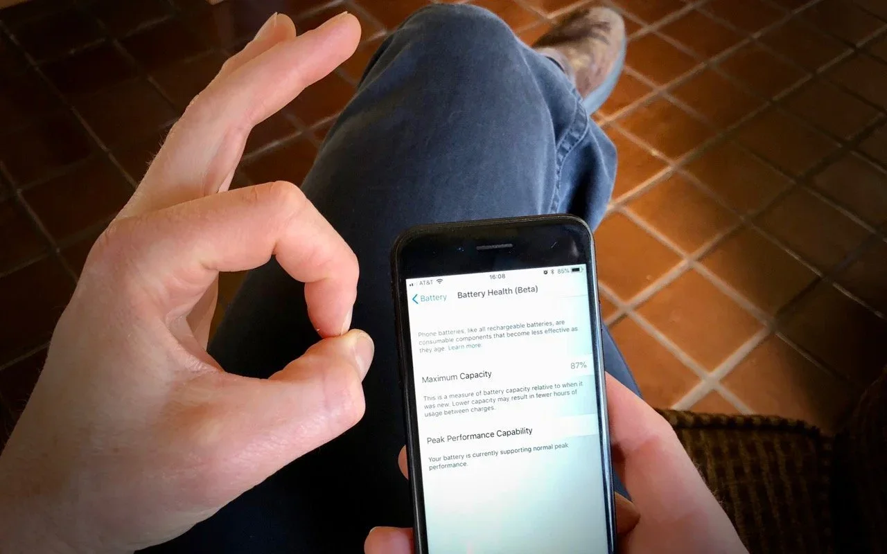 A hand making an OK gesture beside an iPhone battery-health screen